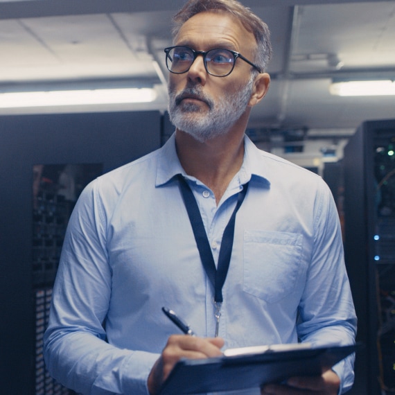 IT specialist inspects server racks in a data center while taking notes on a clipboard.