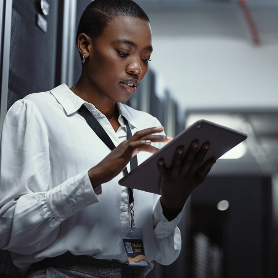 IT specialist reviews system data on a tablet while standing beside server racks.