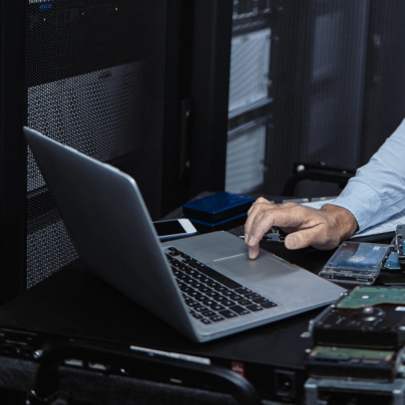 IT specialist analyzes server data on a laptop beside racks.