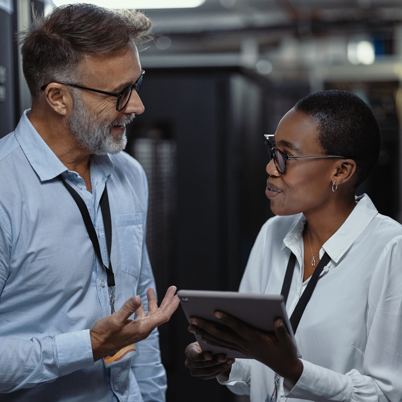 Two IT specialists discuss system updates while reviewing a tablet in a data center aisle.