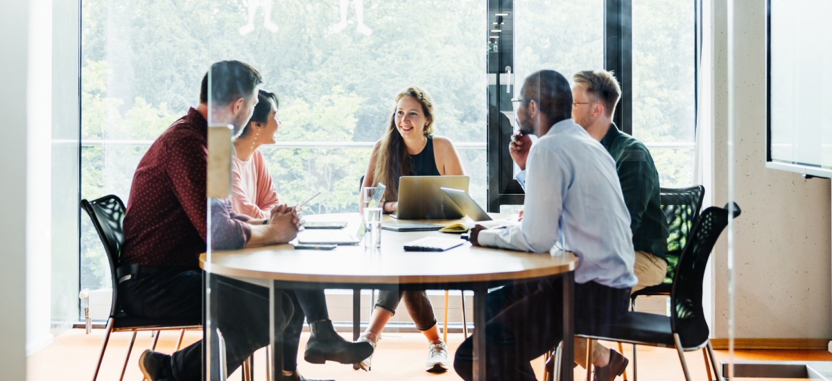 Team members collaborate around a conference table during a meeting in a glass-walled office.