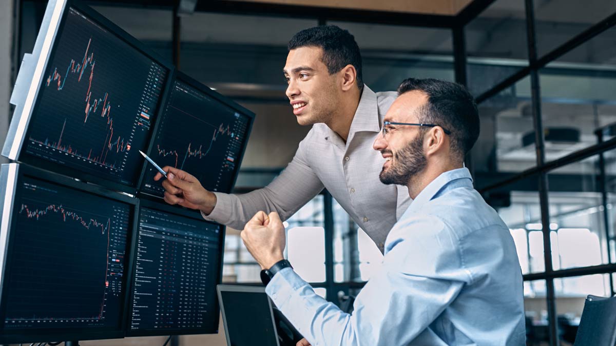 Two male professionals collaborate in front of multiple computer monitors displaying financial charts and data tables. One man points at a screen with a pen while the other raises his fist in a gesture of success. The office setting features large windows and a modern, open-plan workspace. The screens show fluctuating line graphs and numerical information, suggesting active trading or market analysis.