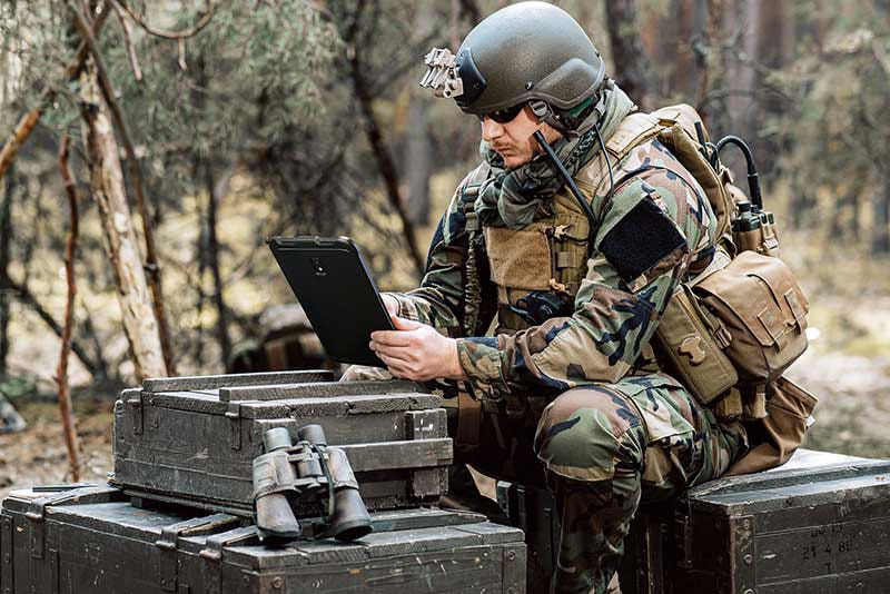 Military personnel using rugged government-issued tablet for secure data access in a remote forest operation, showcasing advanced government technology solutions for field communication and tactical planning.