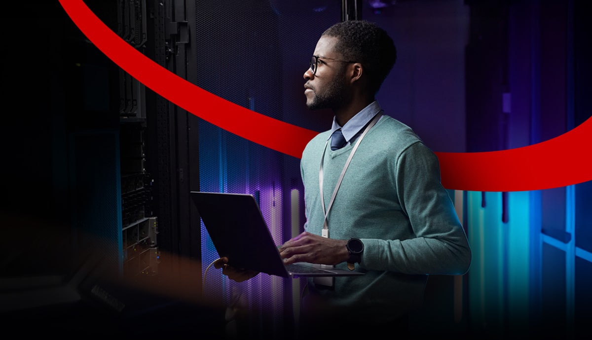 Man with laptop in server room, wearing glasses and a lanyard, with colorful lights and a red graphic overlay.