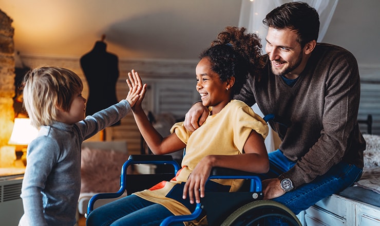 Young girl in a wheelchair high fiving a youg boy with adult in background. 