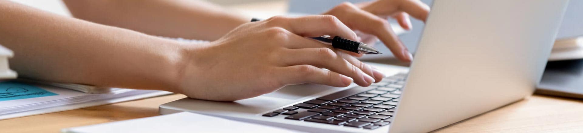 Person at desk, hands typing on laptop with pen in hand