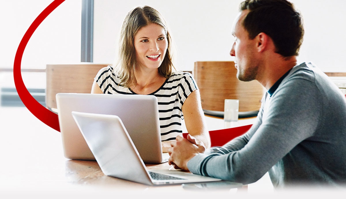 Two professionals at computers discussing work, with a red orbit graphic behind them.