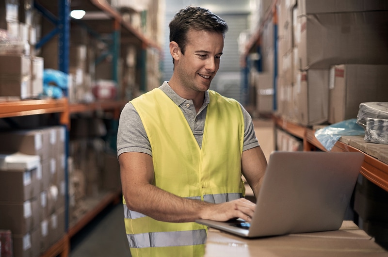 Man in safety vest using laptop in warehouse aisle.