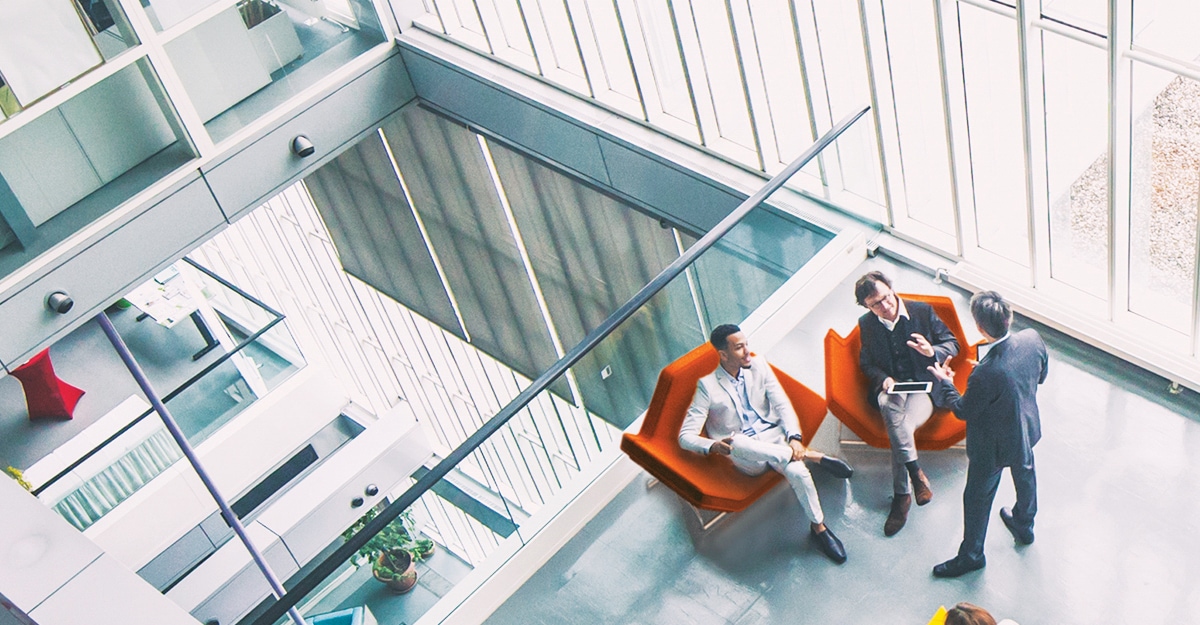 3 People doing a discussion whilst sitting near an indoor balcony, 2 people sitting on chairs with tablets and another person standing.