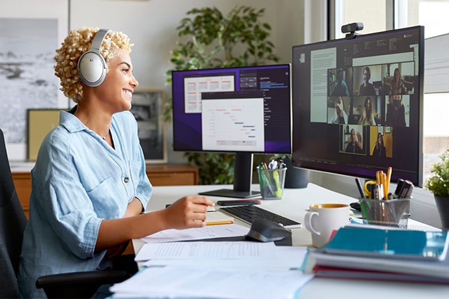 Cheerful businesswoman holding document during video conference with colleagues. Smiling blond Afro female professional is working at home office. She is wearing wireless headphones.